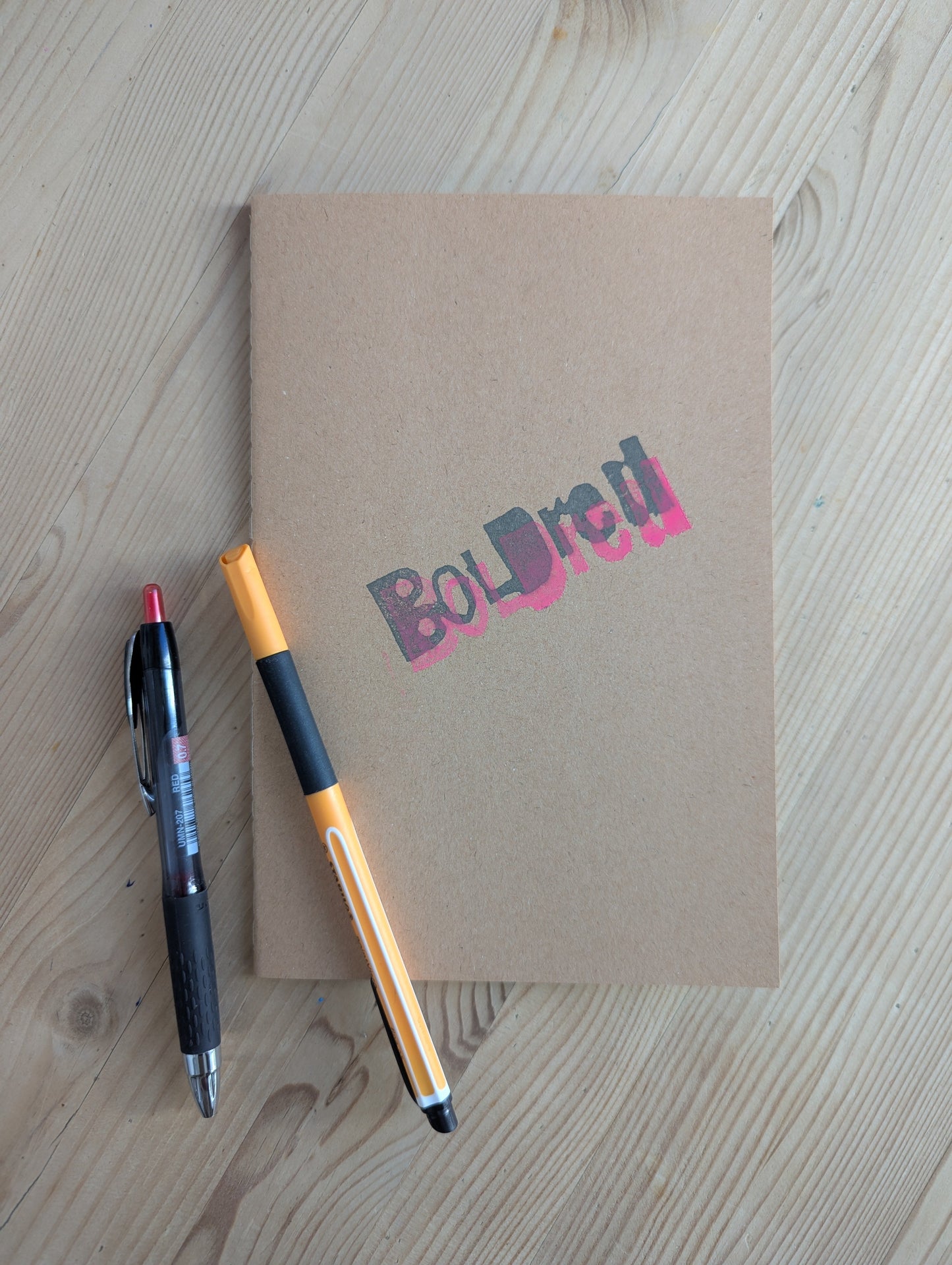 Brown A5 Notebook with brown blank paper with the BOLDred stamp in black and pink places on top of each other. 2 pens are placed next and on top of the notebook to give a visual representation of what a Notebook is for. The objects are all placed on a wooden table.