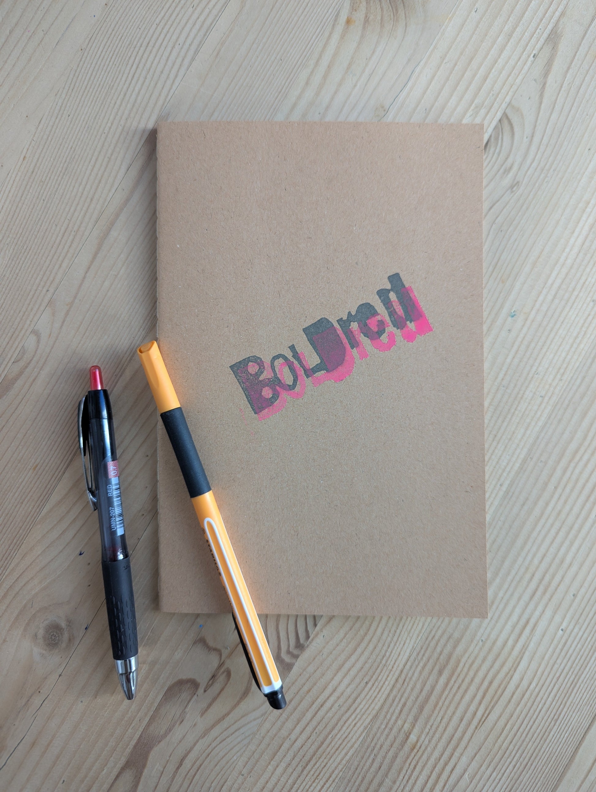 Brown A5 Notebook with brown blank paper with the BOLDred stamp in black and pink places on top of each other. 2 pens are placed next and on top of the notebook to give a visual representation of what a Notebook is for. The objects are all placed on a wooden table.